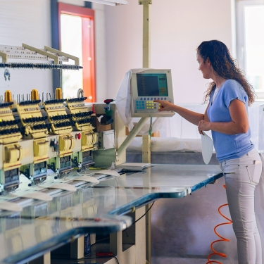 A woman operates an industrial embroidery machine, pressing buttons on a control panel in a well-lit workspace. Sheets of fabric are positioned under the machine’s needles.