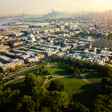 Aerial view of a large city park with green lawns and trees in the foreground, surrounded by dense urban buildings and a distant skyline under a hazy sky.