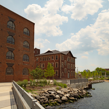 Red brick industrial buildings sit beside a river, surrounded by trees and rocks along the water’s edge. The sky is partly cloudy, and the area appears calm and well-maintained.