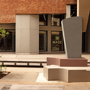 A modern outdoor courtyard with abstract concrete benches and a geometric sculpture, set against a brick building with large windows. One window displays the words MADE in bold letters.