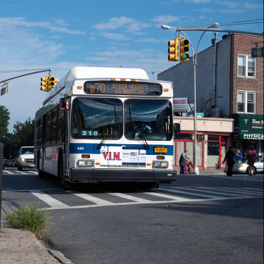 A city bus labeled B70 to Dyker Heights approaches a crosswalk at an intersection on a sunny day, with buildings, traffic lights, and pedestrians in the background.