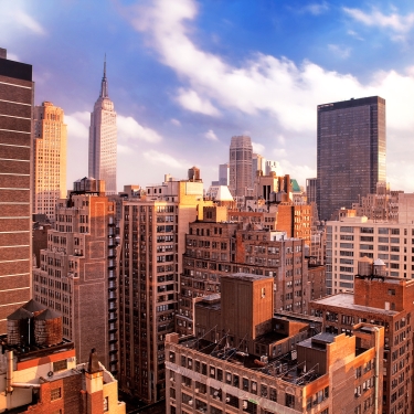 A cityscape featuring tall buildings and skyscrapers under a partly cloudy sky, with the Empire State Building visible in the background.