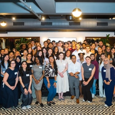 A large, diverse group of people poses together and smiles for a group photo in a brightly lit, modern indoor space with patterned flooring and plants in the background.