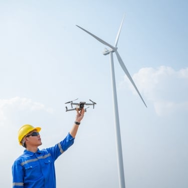 A person in a yellow hard hat and blue workwear holds a drone up toward the sky, standing near a large wind turbine on a clear day.