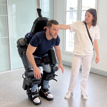 A man wearing a robotic exoskeleton suit bends forward while a woman in white medical attire assists him in a bright room with large windows.