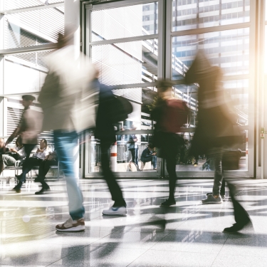 People walking quickly through a bright, modern building lobby with large windows, glass doors, and reflections on the floor. The image is slightly blurred, capturing a sense of motion and bustling activity.