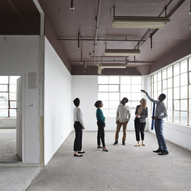 Five people stand in an empty, sunlit office space with large windows. One person gestures upward while others listen, suggesting a discussion or tour of the space. The room has unfinished walls and exposed pipes.