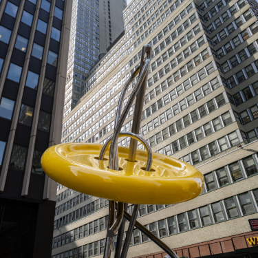 A modern outdoor sculpture featuring a shiny yellow ring intersected by metallic rods, set against a backdrop of tall, glass and concrete office buildings in New York City.