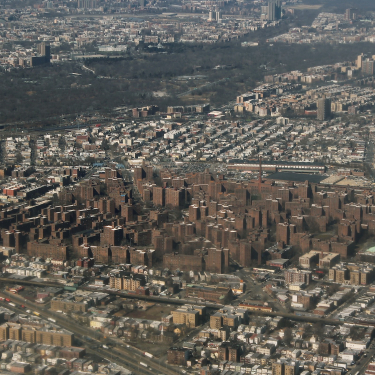 Aerial view of the densely populated Parkchester, Bronx neighborhood with numerous brown apartment buildings, roads, and patches of greenery, surrounded by more residential areas and a large wooded park.
