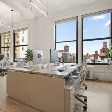 Modern, bright office space with large windows, several desks with computers, ergonomic chairs, and a view of city buildings and water towers outside. Indoor plants add greenery to the clean, minimal decor.