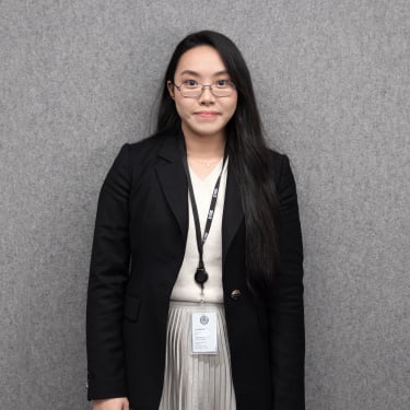 Crystal Ng with long dark hair, glasses, and a black blazer stands in front of a gray background, wearing a lanyard and ID badge. She is looking at the camera with a neutral expression.