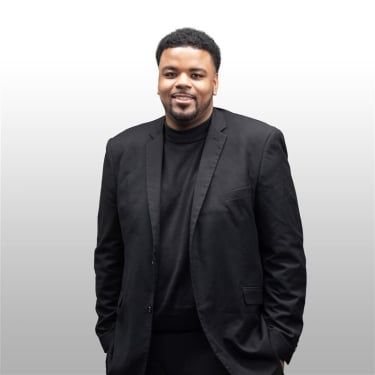 Stephen Jackson wearing a black suit and black sweater stands against a light gray background, smiling with his hands in his pockets.