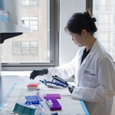 A scientist in a white lab coat and black gloves works with lab equipment and test tubes at a bench near large windows in a laboratory setting.