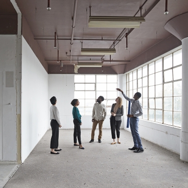 Five people stand in an empty, unfurnished real estate office with large windows, discussing or viewing the space. One person gestures upwards as others listen, suggesting a tour of the property.