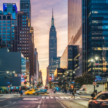 Busy New York City street at sunset with the Empire State Building centered in the background, surrounded by tall buildings, moving cars, taxis, and blurred pedestrians crossing the street.