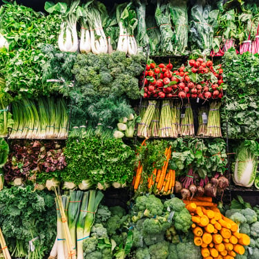 A colorful display of fresh vegetables, including leafy greens, celery, broccoli, carrots, beets, and bok choy, neatly arranged in rows on supermarket shelves.
