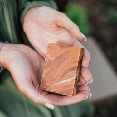 A person wearing a green dress holds a rectangular bar of marbled brown soap with both hands, displaying it outdoors.