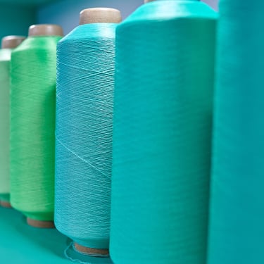 Spools of thread in various shades of blue and green are arranged in a row, with the focus on the nearest spools and the background softly blurred.