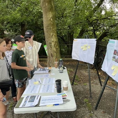 Four people stand by a table with documents and markers, looking at large charts and photos displayed on easels outdoors in a leafy park setting.