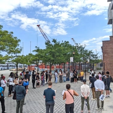 A diverse group of people stands in a semi-circle on a cobblestone plaza near a waterfront, listening to a speaker. There are trees, a brick building, and construction cranes in the background under a partly cloudy sky.