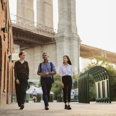 Three people walk and talk together on a city sidewalk near a brick building and a bridge, with trees and a bike rack visible in the background. The scene is bright and appears to be taken during the day.