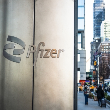 Pedestrians on the sidewalk of a street in Midtown Manhattan, passing Pfizer's headquarters building, with the company name in close-up on an external wall of the building.