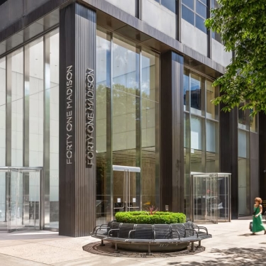 The image shows the glass entrance of the Forty One Madison building, with people walking on the sidewalk nearby and a round bench with greenery in front. Trees partially frame the right side of the scene.