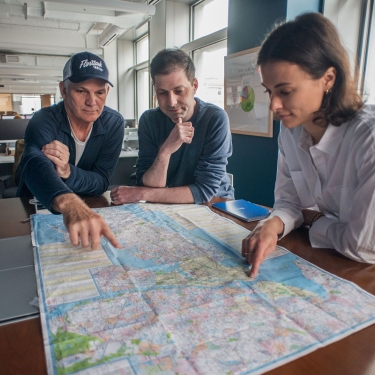 Three people sit at a table in an office, examining and pointing at locations on a large map spread out in front of them. A blue hard hat sits nearby, and large windows let in natural light.