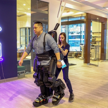 A man wearing a robotic exoskeleton suit is assisted by a woman in a modern indoor space, with a neon sign of a walking figure on the wall and large windows showing cars outside.