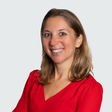 Woman with light brown hair, Rachel Friedman, wearing a red long-sleeve dress stands with arms crossed, smiling at the camera against a plain light gray background.