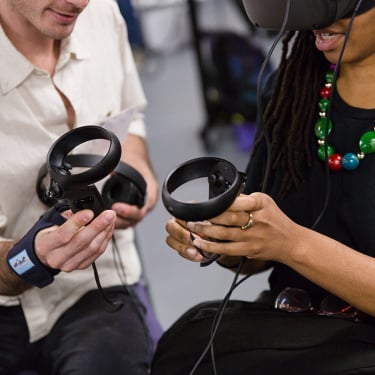 Two people hold virtual reality controllers; one person wears a VR headset and colorful beaded necklace, while the other person, in a light shirt, helps adjust the device. Their faces are partially out of frame.