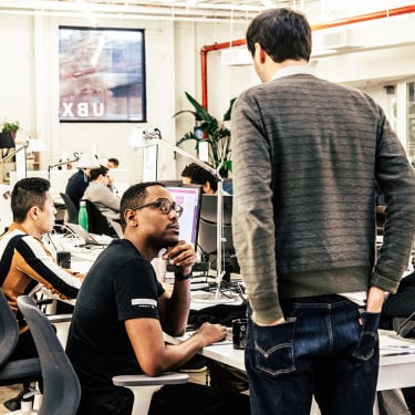 Two men converse at a busy, modern office workspace; one is seated at a computer while the other stands beside him. Other employees work at desks in the background with monitors and office equipment visible.
