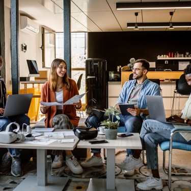 Four people sitting in a modern office, engaged in a casual meeting. They are using laptops and holding papers. The setting includes a coffee table, plants, and a kitchen area in the background, creating a relaxed atmosphere.