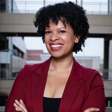 Haizel McIntyre with curly hair, wearing a maroon blazer and black top, stands smiling with arms crossed in an indoor modern building with large windows in the background.