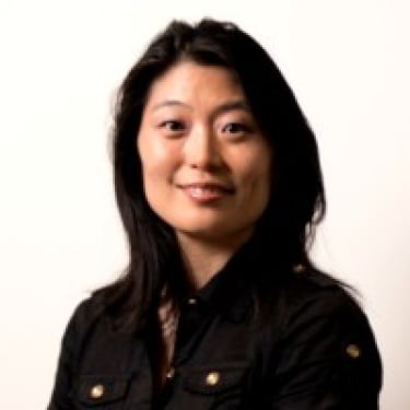 Jane Shin with long black hair wearing a black button-up shirt smiles at the camera against a plain light background.
