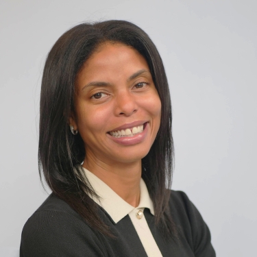 Saira Patel with straight dark hair and medium skin tone smiles at the camera. She is wearing a black jacket over a light-colored collared shirt, set against a plain light background.