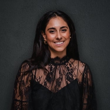 Valeria Mendoza with long dark hair smiles at the camera, wearing a sheer black lace top with a high neckline and gold hoop earrings, standing against a dark textured background.