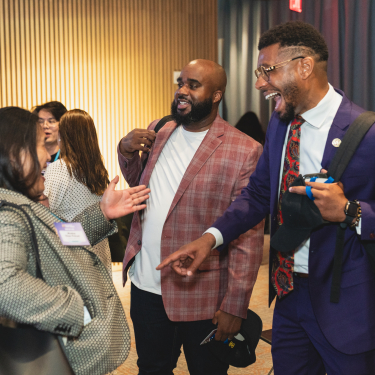 Three people stand together indoors, smiling and laughing as they converse. Two men with backpacks face a woman wearing a patterned coat. Other people are visible in the background near wooden wall panels.