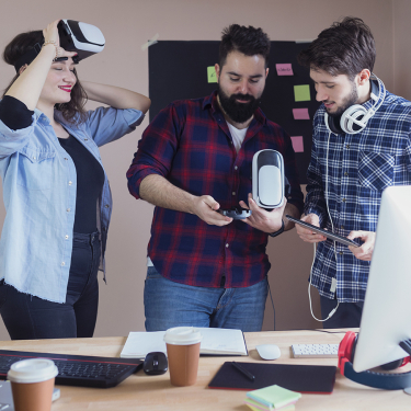 Three young adults in a modern office interact with virtual reality headsets; one wears a headset, while the other two examine another headset and a tablet. Sticky notes and coffee cups are visible on the desk.