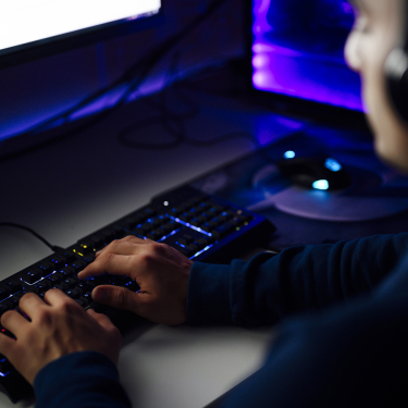 A person types on a backlit keyboard in a dimly lit room, with a computer monitor and a glowing mouse visible on the desk. The scene suggests focused computer work or gaming.