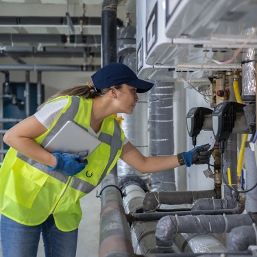 A person in a neon safety vest and cap inspects industrial equipment in a mechanical room, holding a tablet and wearing blue gloves. Pipes and control panels are visible in the background.