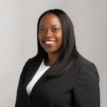 Patricia Jacques with long straight black hair, wearing a black blazer over a white top, smiles at the camera against a plain light gray background.