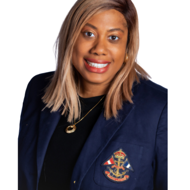 Sarah Perkins with straight, shoulder-length blonde hair smiles at the camera. She is wearing a navy blazer with an embroidered crest, a black top, and a gold necklace, against a white background.