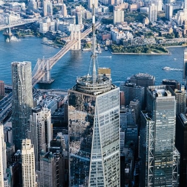 Aerial view of Lower Manhattan skyscrapers with the Brooklyn and Manhattan Bridges crossing the East River, and Brooklyn visible in the background on a clear day.