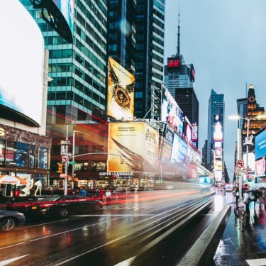 A busy Times Square street scene in New York City at dusk, with bright billboards, blurred car lights, and crowds of people walking in the rain. Skyscrapers surround the bustling urban intersection.