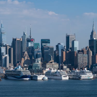 New York City skyline with tall skyscrapers, including the Empire State Building, seen from across the Hudson River with several large cruise ships docked at the Manhattan Cruise Terminal under a blue sky.