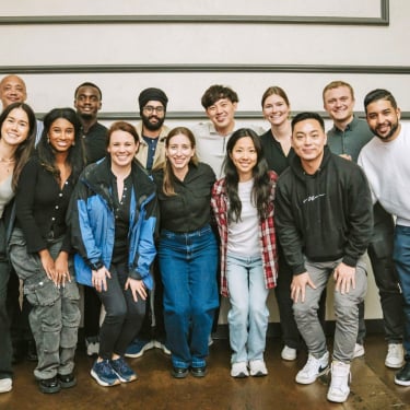 A group of thirteen people stand and kneel together indoors, smiling at the camera. They are dressed casually, and the setting appears to be a social or professional gathering.