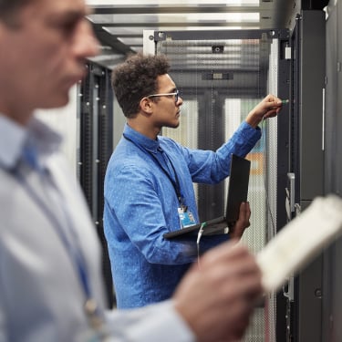 Two IT professionals work in a server room; one in focus adjusts hardware on a server rack while holding a laptop, while the other, blurred in the foreground, inspects a device or document.