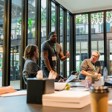 A man stands and speaks to two seated colleagues in a modern glass-walled office, engaging in a discussion. Papers, notebooks, a coffee cup, and water bottles are on the table in front of them.