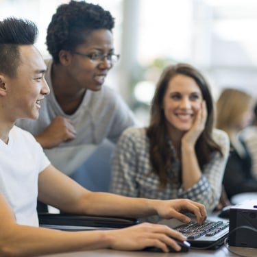 Three young adults work together at a computer. One person types while the others watch and smile, appearing engaged and collaborative in a bright, modern setting.